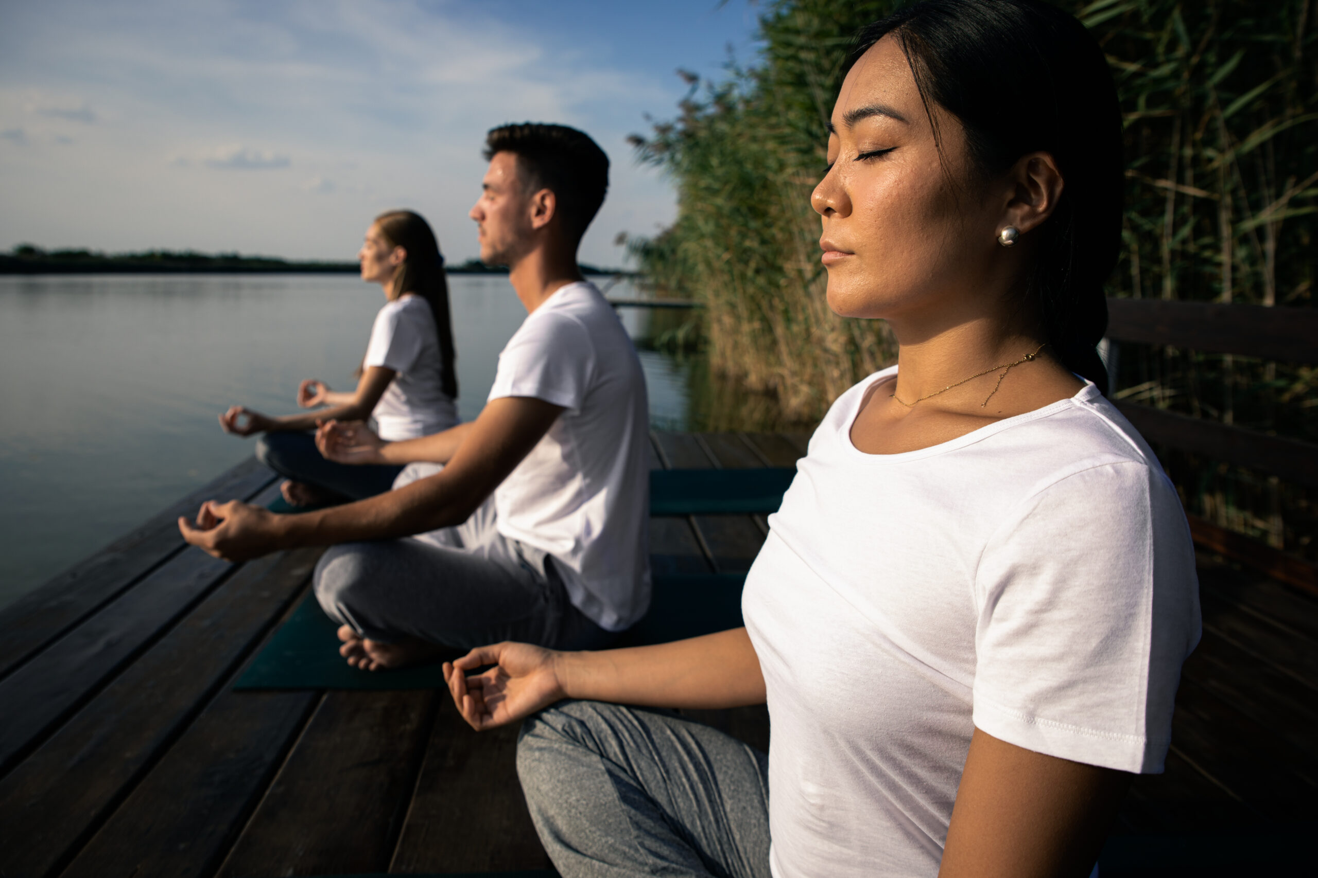 Three people practicing mindful breathing meditation by a lake, sitting calmly outdoors to reduce stress and improve focus